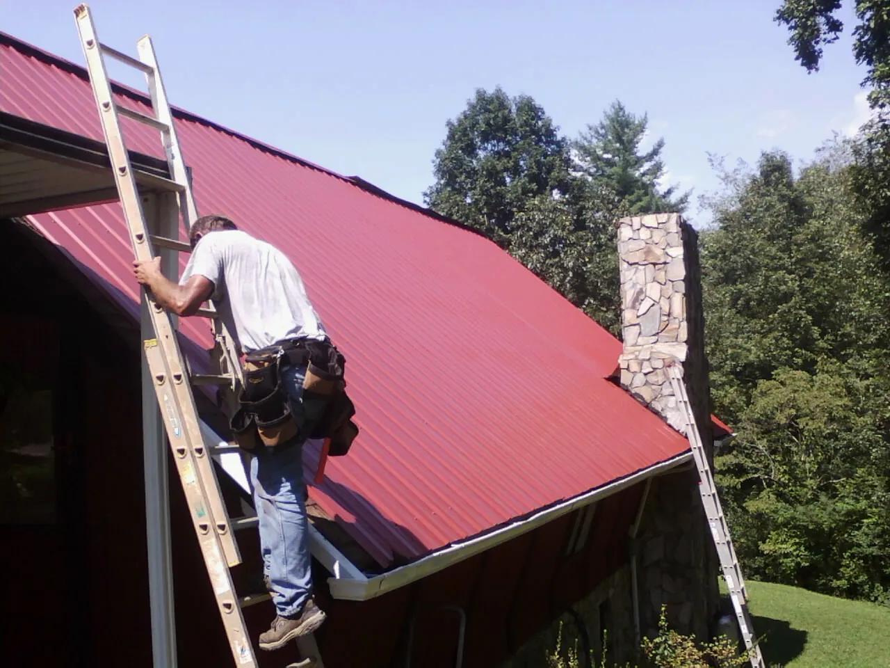 Roofer on a ladder beside a red metal roof and stone chimney — Soffit & Fascia Repair by Roofing Brothers in Blakesburg